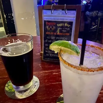 A pint of dark beer and a cocktail with a lime wedge and chili rim sit on a bar table with a happy hour menu in the background.