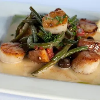 Seared scallops with green beans, olives, diced tomatoes, and fresh herbs served in a light cream sauce on a square white plate with a lemon wedge.