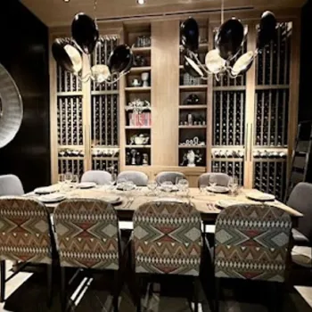 A dining room with a long table set for eight, patterned chairs, wall shelves with wine bottles and dishes, hanging black lights, and a decorative round mirror and textile art on the walls.