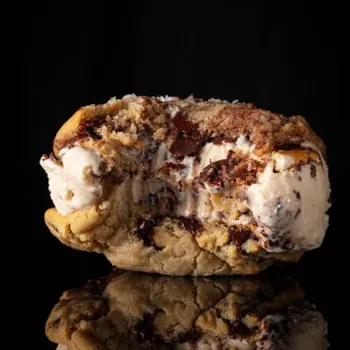 A bitten ice cream sandwich made with chocolate chip cookies and vanilla ice cream, shown against a black background with its reflection visible below.