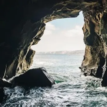 View from inside a rocky sea cave looking out toward the ocean, with waves crashing against rocks and a clear sky visible through the cave opening.