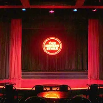A comedy club stage with red neon lights, curtains, and round tables with chairs in the foreground.