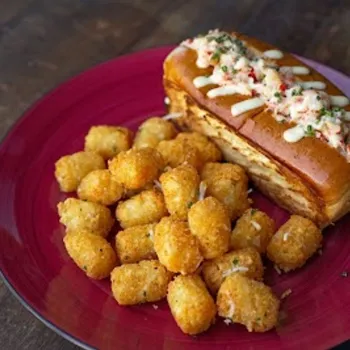 A plate with a lobster roll topped with herbs and tater tots, served on a red dish on a wooden surface.