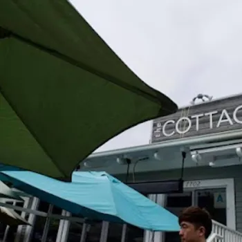 Outdoor patio area with green and blue umbrellas in front of a gray building with a sign that reads "The Cottage." A person is partially visible at the bottom.