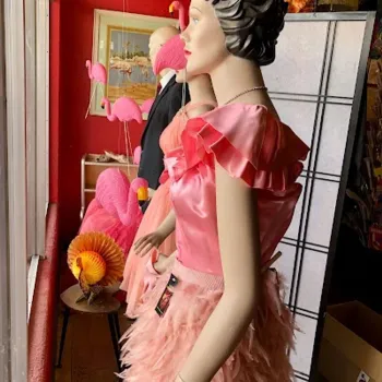 A mannequin dressed in a pink ruffled top and feathered skirt stands by a window, surrounded by flamingo decorations and a paper turkey in a colorful store display.