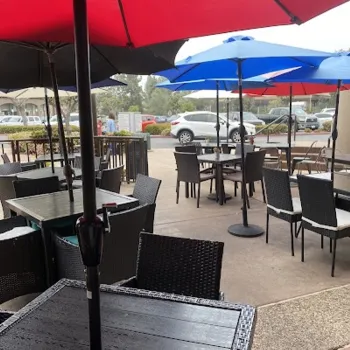 Outdoor patio dining area with black tables and chairs under large red and blue umbrellas; parked cars and a sidewalk are visible in the background.