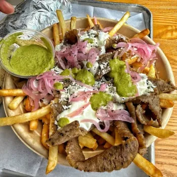 A bowl of French fries topped with sliced gyro meat, pickled onions, white sauce, green sauce, and crumbled cheese, served with a side of green sauce on a wooden table.