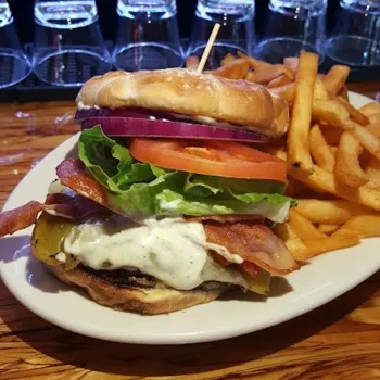 A cheeseburger with bacon, lettuce, tomato, and red onion on a bun, served with French fries on a white plate at a wooden counter.