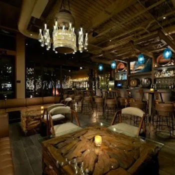 Dimly lit lounge with wooden furniture, a chandelier, leather seating, a bar area with high stools, and decorative lighting.