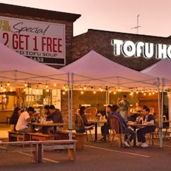 People dine outdoors under white canopy tents at evening, in front of a restaurant called "Tofu House" with string lights and a "Buy 2 Get 1 Free" sign visible.