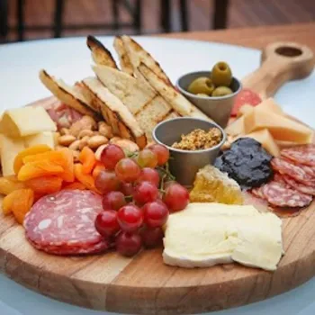A wooden board with assorted cheeses, cured meats, sliced bread, olives, nuts, grapes, dried apricots, mustard, and spreads arranged neatly.