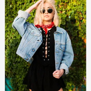 Woman wearing sunglasses, a red bandana, a denim jacket, and a black dress poses confidently in front of a leafy green background.