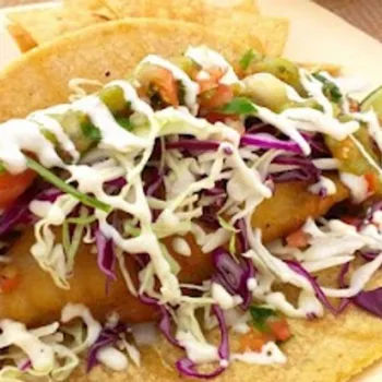A fish taco topped with shredded cabbage, salsa, and white sauce is served on a corn tortilla with lime wedges and tortilla chips on the side.
