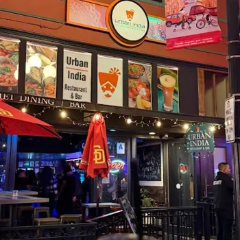 Nighttime view of Urban India Restaurant & Bar exterior with illuminated sign, outdoor seating, red umbrellas, and a close-up of a drink in the foreground.