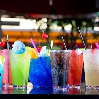 A row of colorful cocktails and soft drinks with straws and garnishes sit on a bar counter at an outdoor venue.