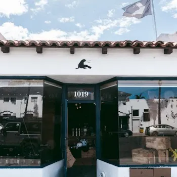 A storefront with an open door, number 1019 above the entrance, a flag on the roof, and various items visible inside. A bench sits outside on the sidewalk.