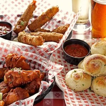 Baskets of fried mozzarella sticks, chicken wings, garlic knots with dipping sauces, and a pint of beer on a red checkered tablecloth.