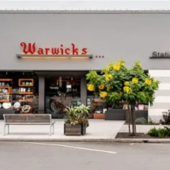 Street view of Warwick's storefront with large display windows, plants, and two small trees with yellow flowers on the sidewalk. Signs read “Stationery”, “Gifts”, and “Books.”.