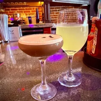 Two cocktails on a bar counter: one is a creamy dark drink in a coupe glass with garnish, the other is a pale yellow drink in a wine glass. Bottles and condiments are in the background.