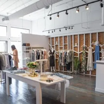 A boutique clothing store interior with racks of clothes, a display table with accessories, and a woman browsing garments near large front windows.