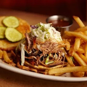 A plate with a pulled pork sandwich topped with coleslaw, sliced pickles on the bun, a side of French fries, and a small cup of barbecue sauce.