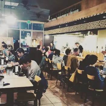 People are seated at tables and the counter in a busy restaurant, eating and talking. The kitchen area and shelves of bottles are visible in the background.