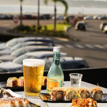 Plates of sushi, a glass of beer, and a bottle of sake on an outdoor table overlooking a parking lot, palm trees, and a waterfront view.