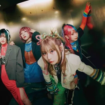 Four young women dressed in colorful, eclectic clothing pose together in a metallic elevator, looking directly at the camera with serious expressions.