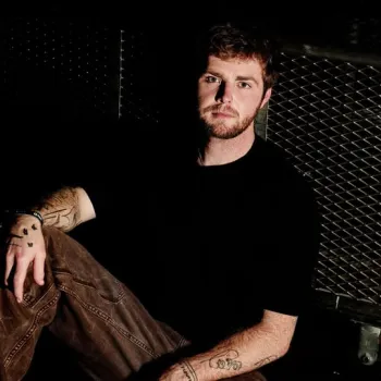 A man with tattoos sits on the floor next to a large metal cart in a dimly lit space, wearing a black t-shirt and brown pants.