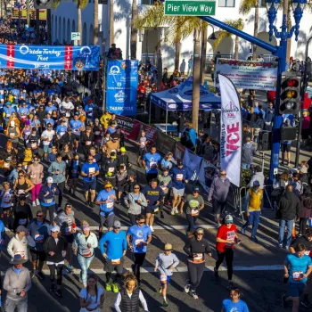 A large crowd of people participate in a road race event, running under a blue event banner on a city street lined with spectators and flags.