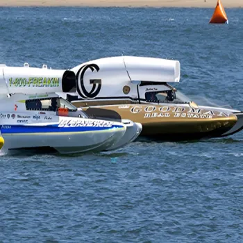 Four colorful hydroplane boats race side by side on the water, creating splashes as they speed toward an orange buoy near a sandy shore.
