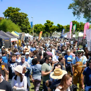 A large crowd of people walks through an outdoor festival or market lined with vendor tents on a sunny day.