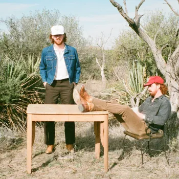 Two men in casual clothes and hats are outdoors by a wooden table; one stands while the other sits with feet propped on the table. Desert plants and trees are in the background.