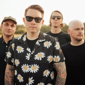 Four men stand outside in a field, all wearing dark sunglasses. The man in front has tattoos and a daisy-patterned shirt. The background is blurred greenery under bright daylight.
