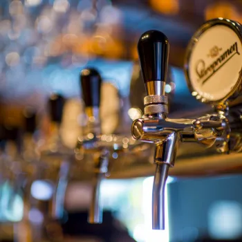 Close-up of beer taps at a bar, with the focus on one tap handle and a blurred background of glassware and lights.