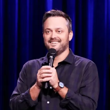 A man stands on stage with a microphone, smiling, in front of dark blue curtains.