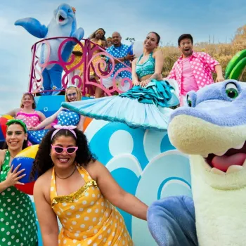 A group of people in bright costumes and animal mascots pose on and around a colorful parade float with beach and ocean decorations.