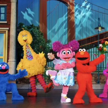 Sesame Street characters, including Big Bird, Elmo, and Cookie Monster, stand on stage in colorful costumes, striking playful poses in front of a city-themed backdrop.