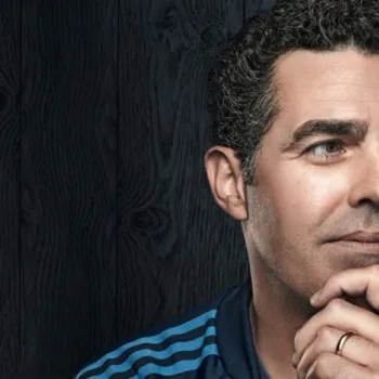 A man with short curly hair and wearing a striped blue jacket looks thoughtfully to the side, resting his chin on his hand, against a dark wooden background.