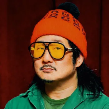 A man with long dark hair, a mustache, and goatee wears oversized yellow-tinted glasses, a red knit beanie, and a green shirt, standing against a dark red background.