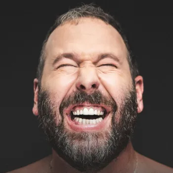 A bearded man with closed eyes smiles widely, showing his teeth, against a dark background.