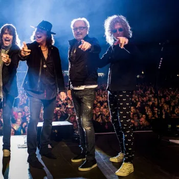 Seven men stand on stage in front of a large crowd, posing for a photo with some pointing toward the camera.