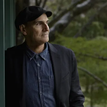 A man in a dark jacket, blue shirt, and black cap stands by a green wall, looking thoughtfully into the distance with trees in the background.