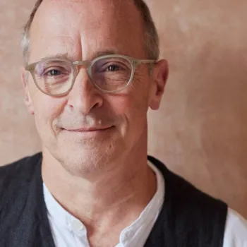 A middle-aged man with short hair and glasses, wearing a white shirt and dark vest, stands in front of a beige textured background.