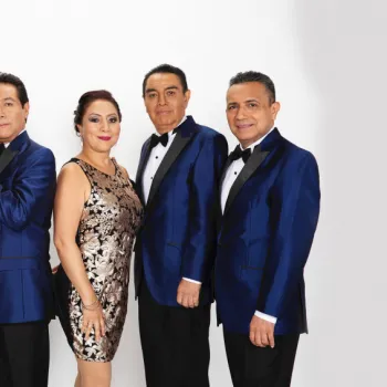 Six adults pose in formal attire against a white background; three men wear blue tuxedos, while two women wear black and gold dresses and one man wears a black tuxedo.