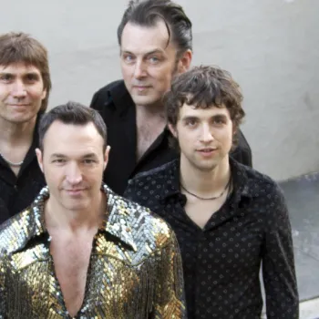 Six men stand together, five in black shirts and one in a gold sequin shirt, posing for a group photo against a plain background.