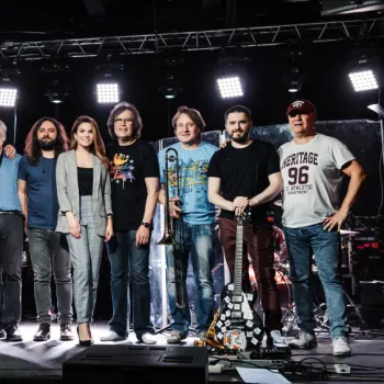 A group of twelve people, some holding musical instruments, stand together on a stage under bright lights, posing for a group photo.