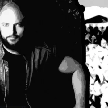 A person with a beard sits on an ornate chair, wearing a leather vest and dark shirt, in a high-contrast black and white image.