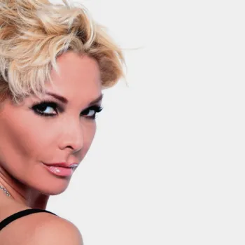 A woman with short, tousled blonde hair and dramatic makeup looks over her shoulder, wearing a black top and a silver necklace against a plain light background.