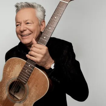 An older man with gray hair smiles while holding an acoustic guitar against a plain light background.